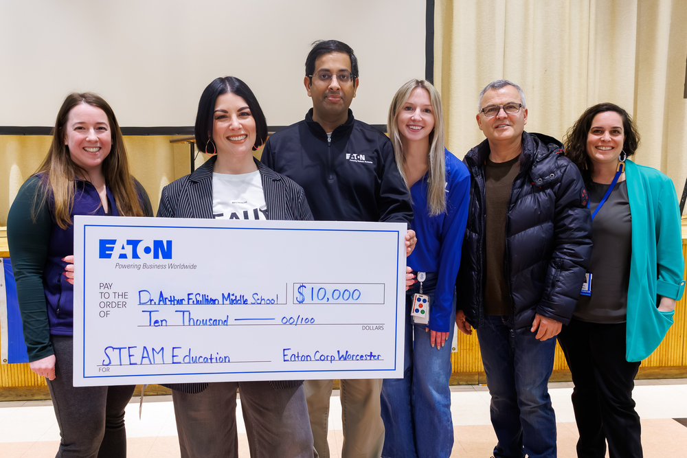 Six adults stand side-by-side, one is holding a check with the value of $10,000.