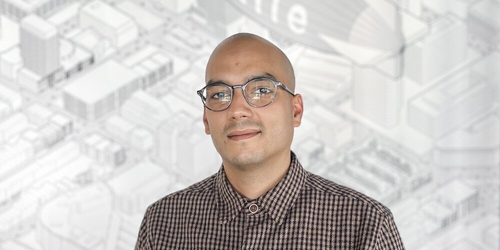 A man with glasses and a bald head smiles gently, with a blurred architectural blueprint in the background.