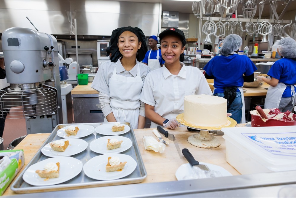 Two smiling bakers pose in a commercial kitchen, surrounded by baked goods.