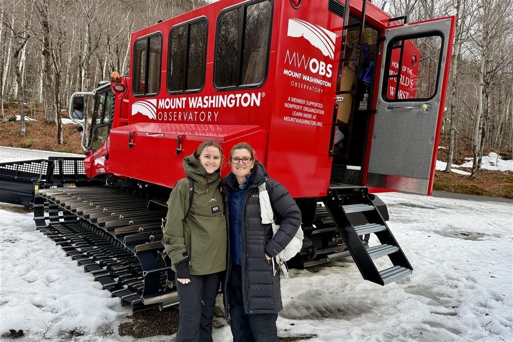 Two people stand in front of a large red Mount Washington Observatory vehicle with tracks.