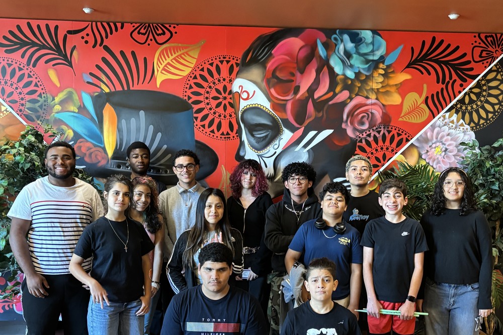 A diverse group of young people pose in front of a colorful mural.