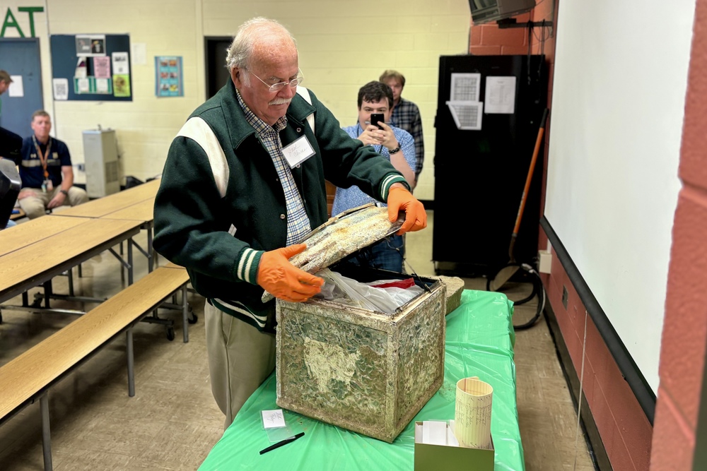 A man in a green jacket opens a weathered box, wearing orange gloves.