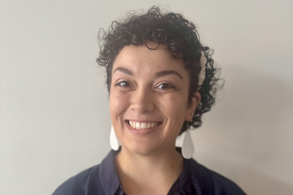 A woman with curly dark hair smiles warmly at the camera, wearing large white earrings and a dark blue shirt.