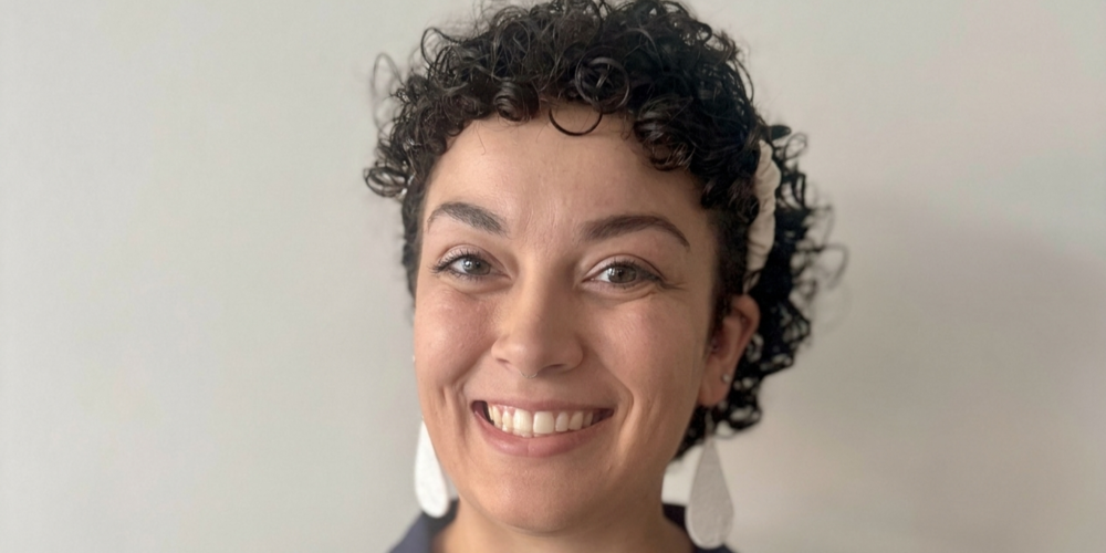 A woman with curly dark hair smiles warmly at the camera, wearing large white earrings and a dark blue shirt.