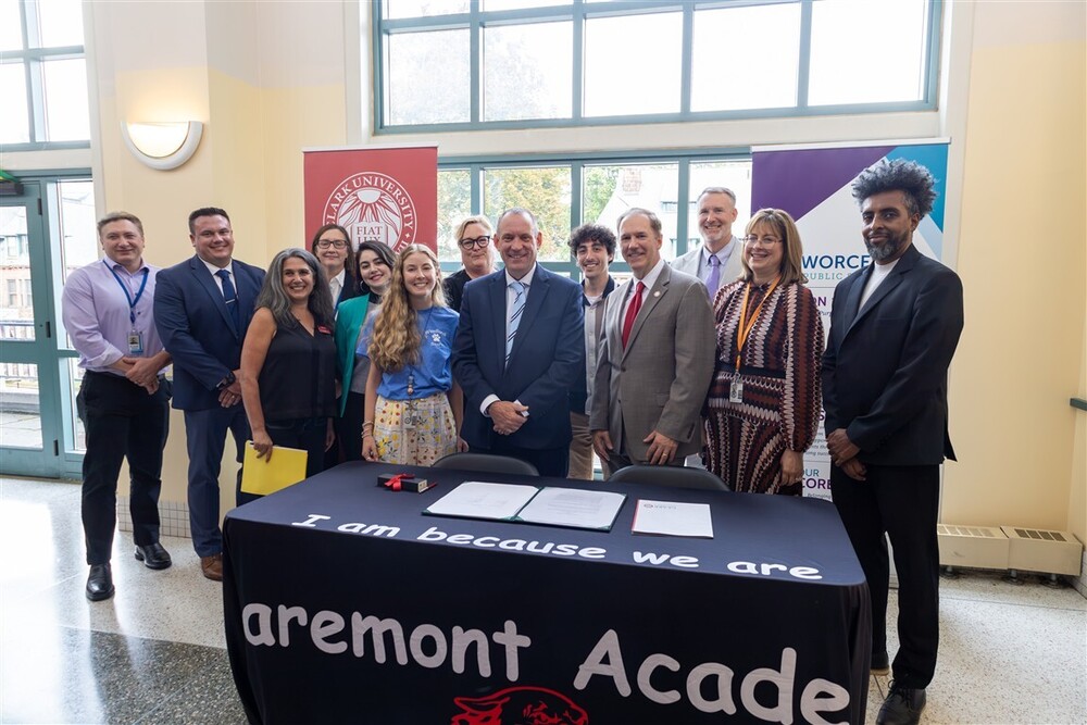 A diverse group of adults poses together behind a table draped with a banner reading "I am because we are Claremont Academy".