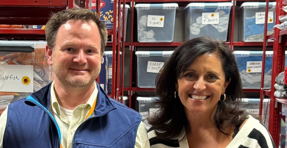 Doherty Memorial High School Principal John Staley and Juniper Outreach Foundation Executive Director Julie McDonald smile at the camera in front of a storage shelf filled with labeled bins of food and essential supplies.