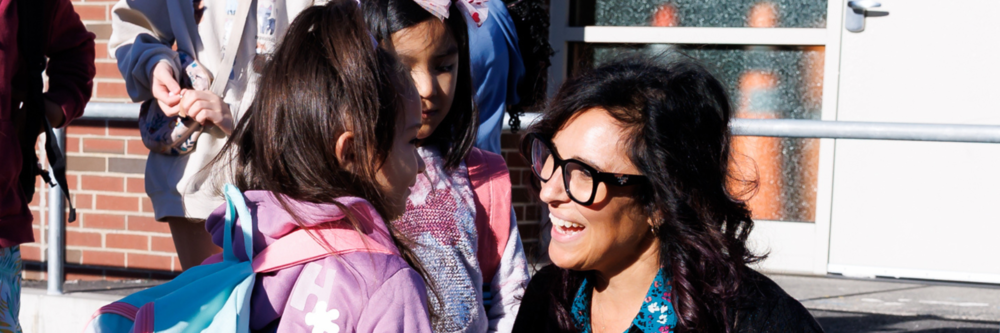 A teacher kneels to speak with a young girl, surrounded by other children, outside a building.