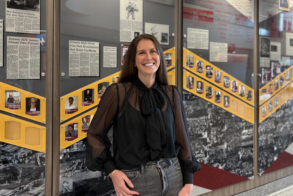 A smiling woman in a black sheer blouse and jeans stands in front of a wall display with framed newspaper clippings and photos.