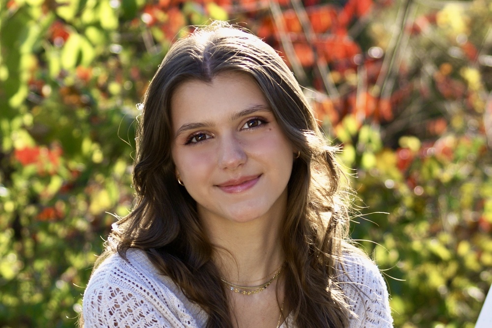 A young woman with long brown hair smiles gently at the camera, with a blurred background of green and orange foliage.