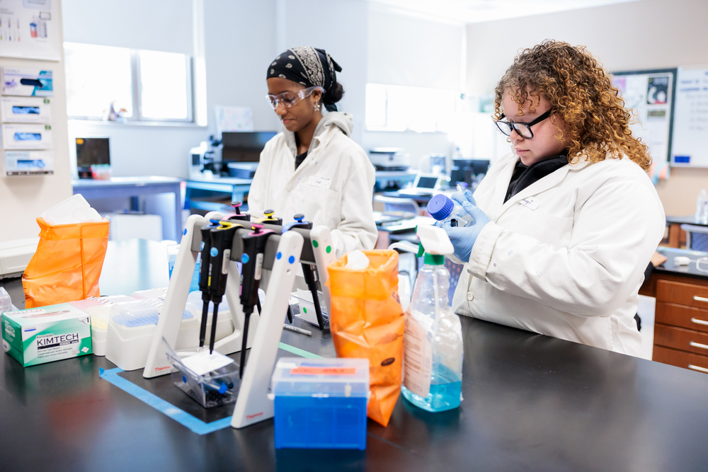 Two students in lab coats work in a brightly lit laboratory setting.