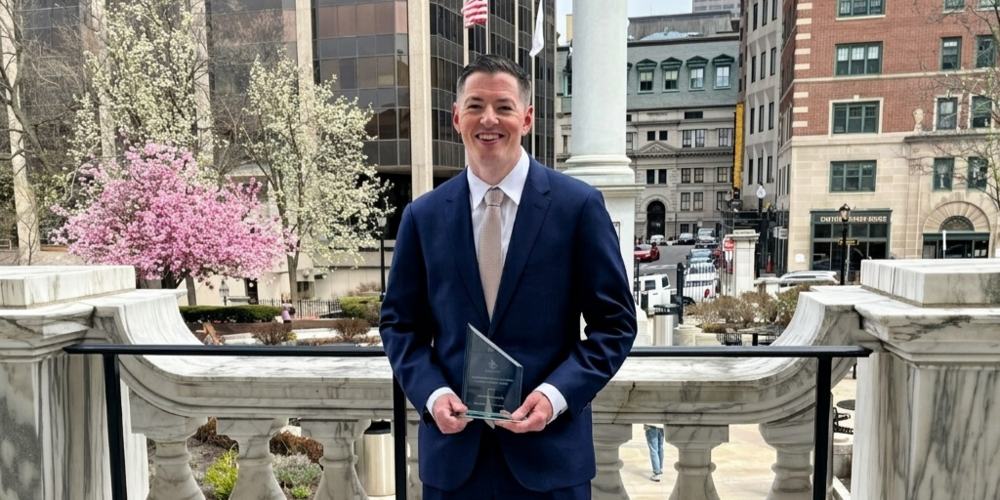 A man in a suit holds a glass award in front of a city backdrop with blooming trees.