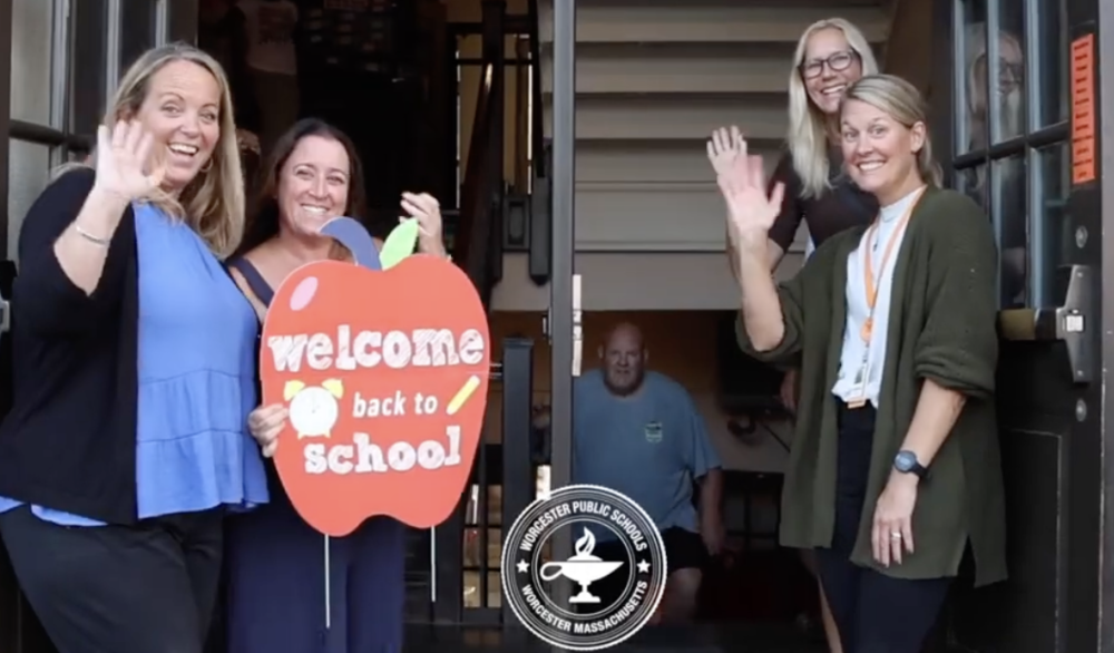 Four women waving and one holding a decorative apple that says "welcome back to school"