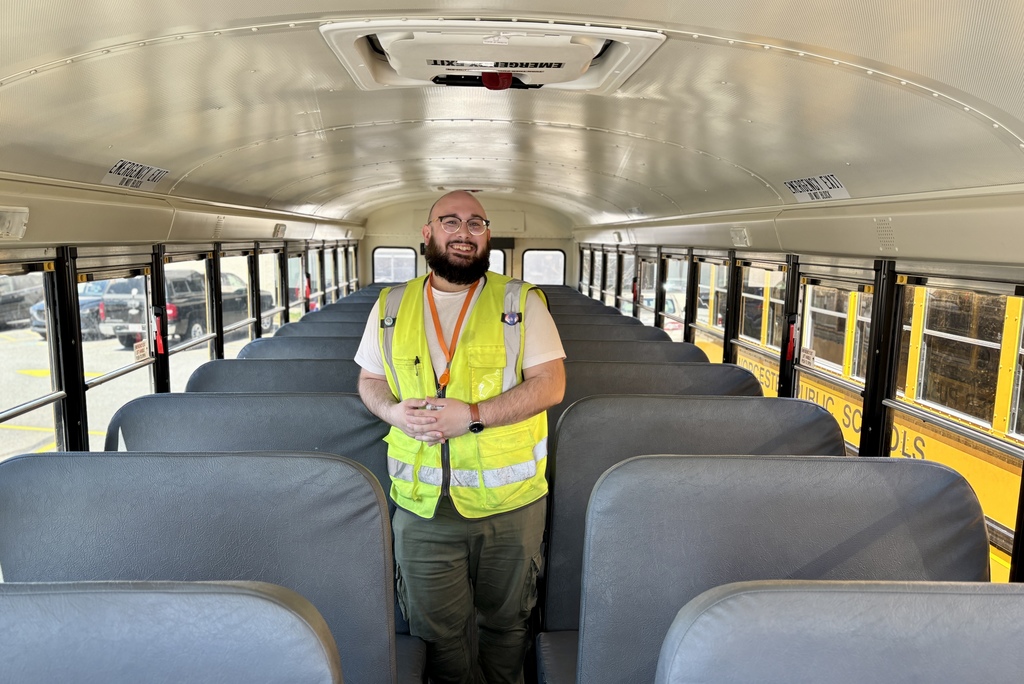 A man in a yellow safety vest stands inside a school bus, smiling at the camera.
