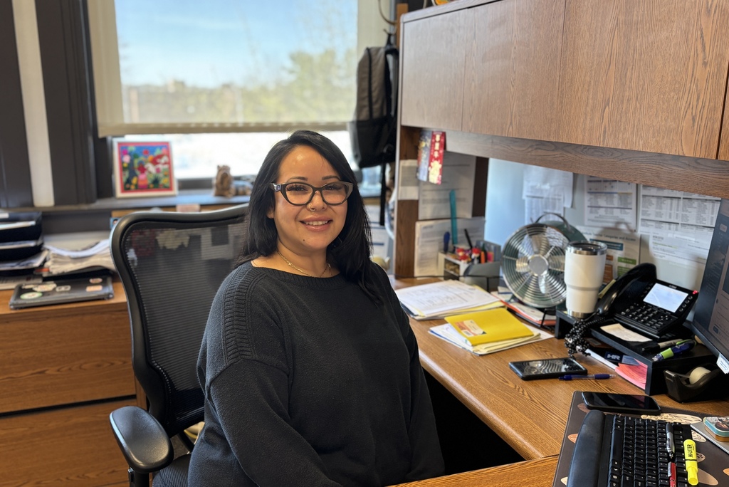A woman with glasses smiles at the camera while sitting at her desk in an office.