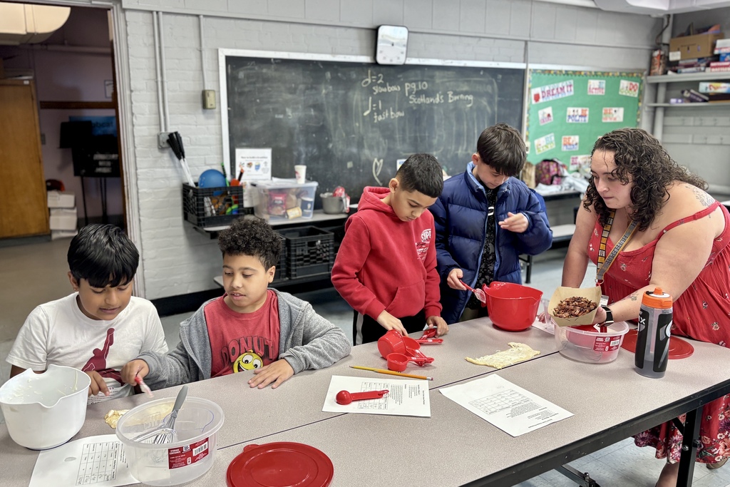 A group of students and a teacher work together in a classroom, using measuring cups and bowls.