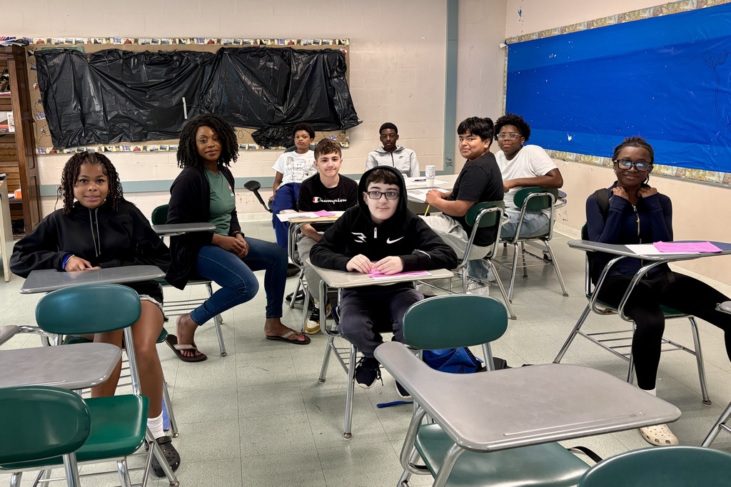 A group of students and their teacher sit together at classroom desks, actively engaged in learning and discussion.