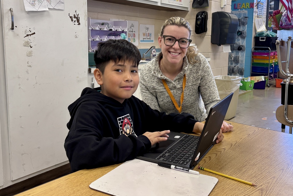 A young student works on a laptop with a teacher in a classroom setting.