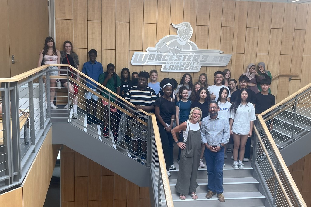 A diverse group of students and two adults pose on a staircase in front of a university logo.