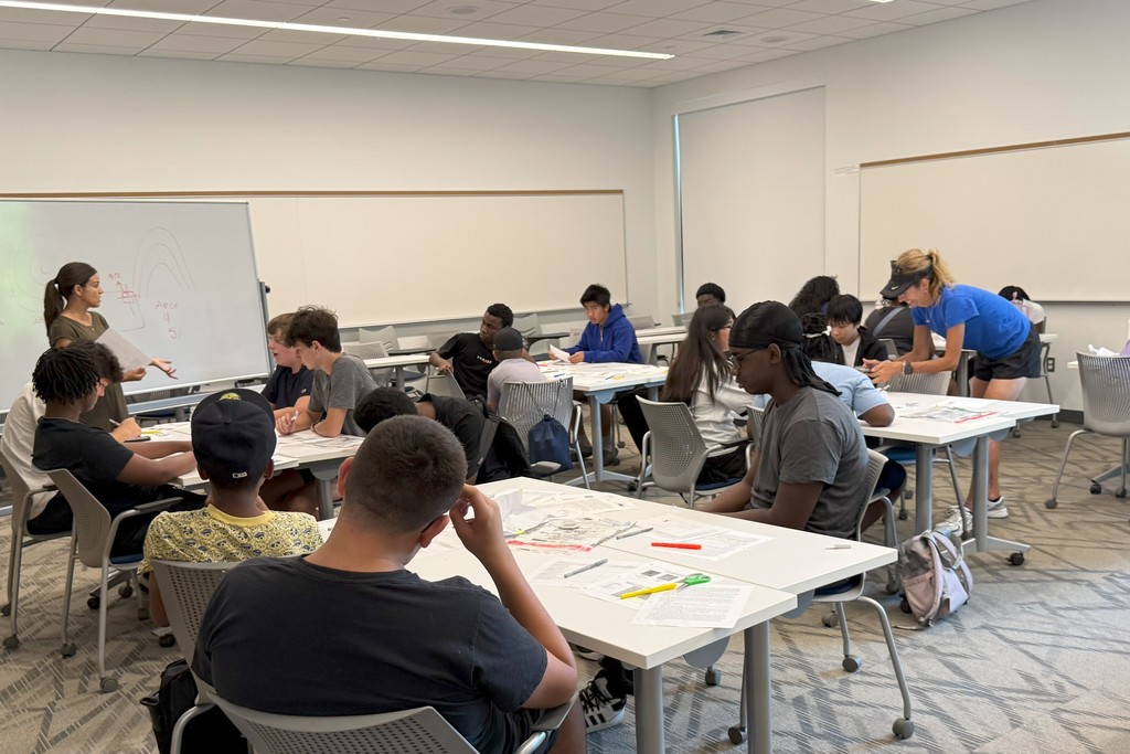 Students work at desks in a classroom setting, with a teacher assisting.
