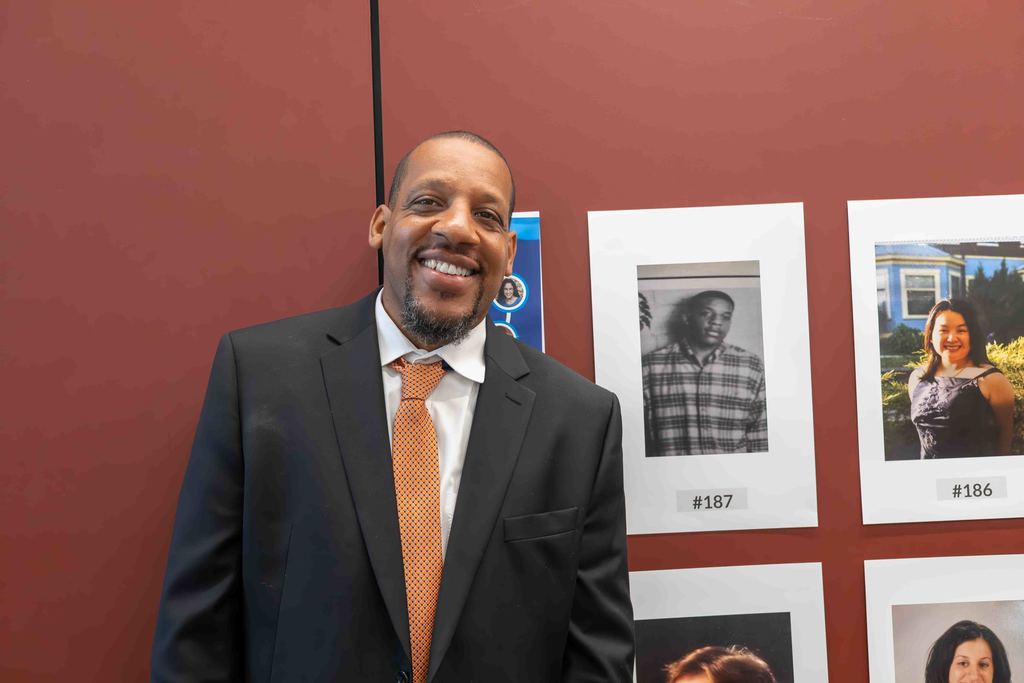 A smiling man in a suit stands in front of a wall with framed photos.