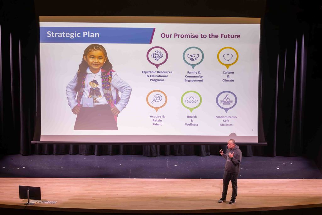 A presenter stands on a stage in front of a screen displaying a strategic plan.