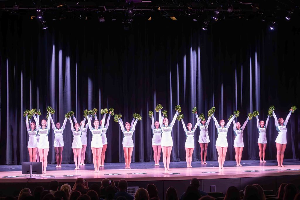 A cheerleading squad performs on stage, holding up green pom-poms.