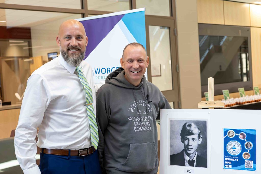 Two men smile, standing near a display with a black and white photo and a sign.