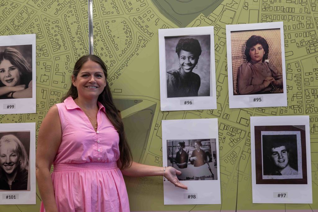 A woman in a pink dress smiles, pointing to a display of framed photos.