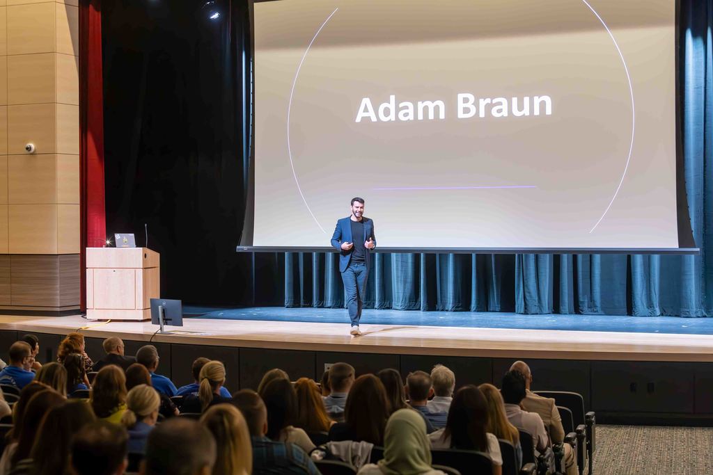 A man in a suit walks on stage in front of an audience, with the name 'Adam Braun' displayed.