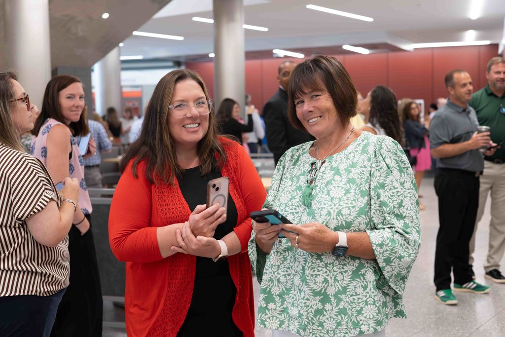 Two women smile while holding phones in a well-lit indoor space.