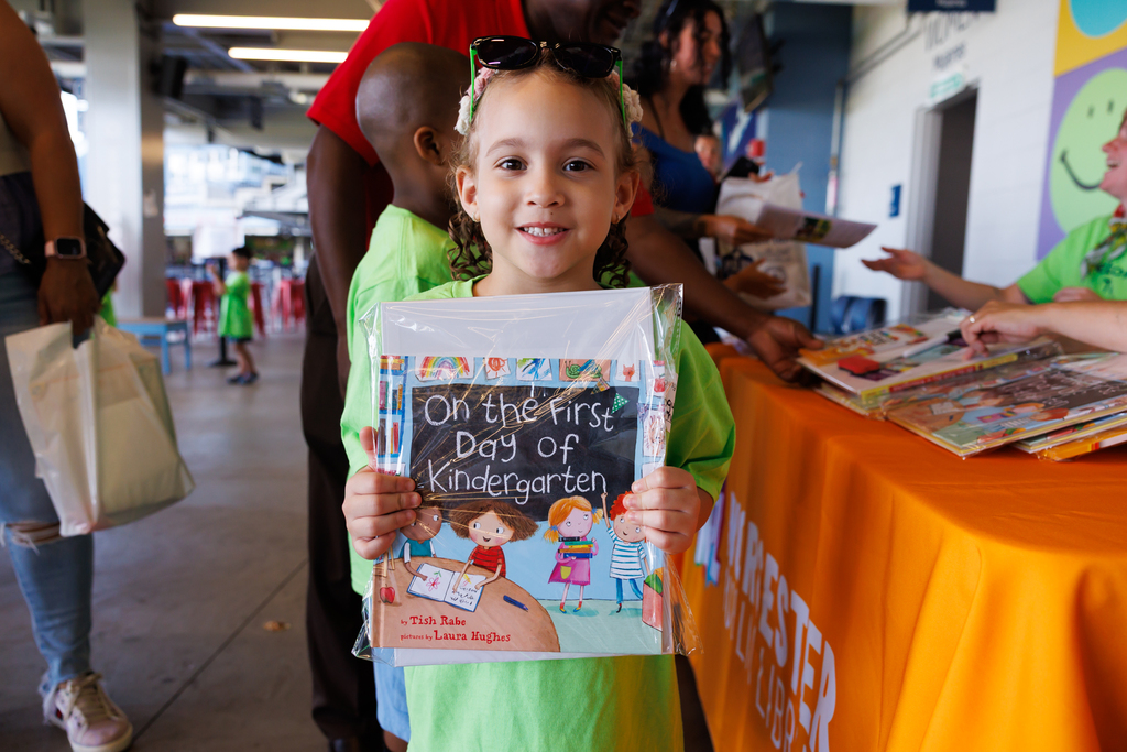 A young girl smiles, holding a book titled 'On the First Day of Kindergarten'.