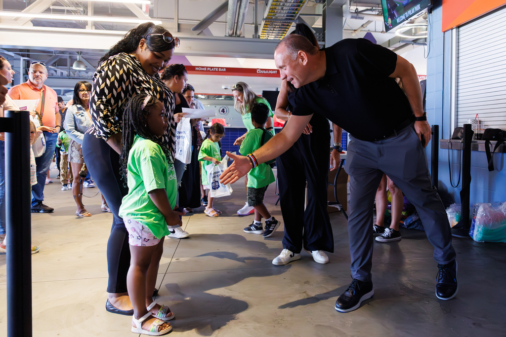 A man extends his hand to greet a young girl in a green shirt, surrounded by a crowd.