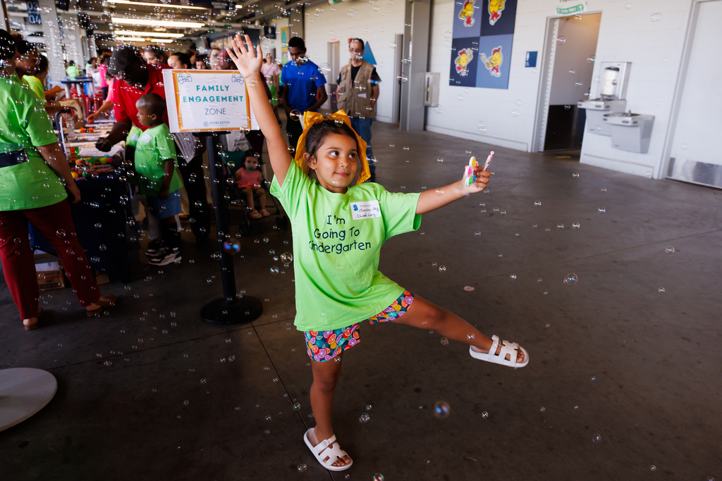 A young girl in a green shirt and colorful shorts plays with bubbles, raising her arms.