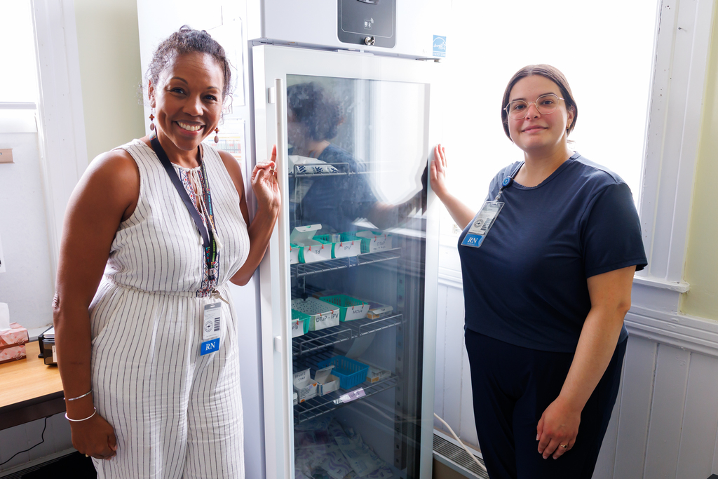 Two nurses stand beside a medical refrigerator, smiling at the camera.