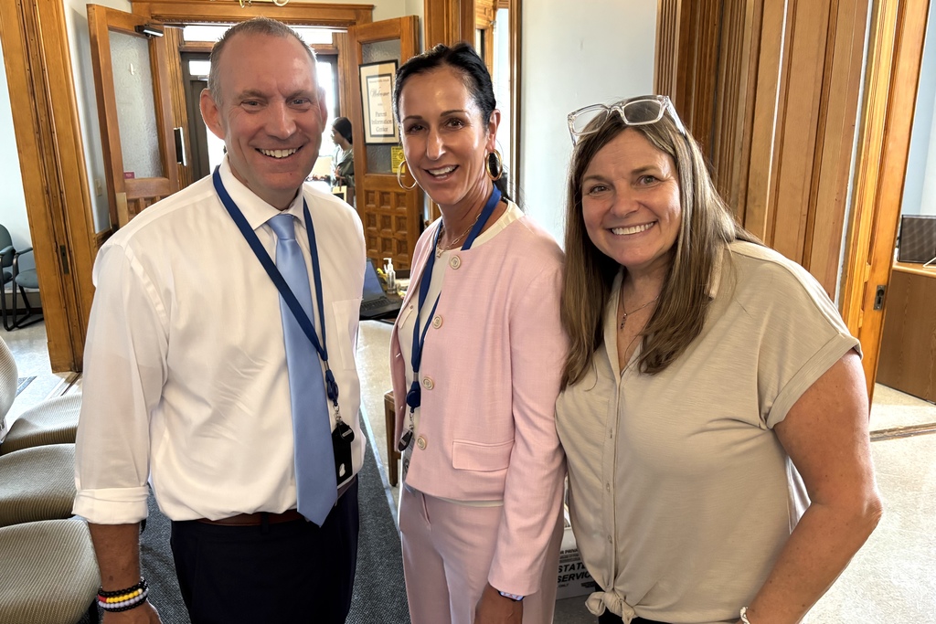 Three smiling people pose for a photo indoors, near wooden doors.