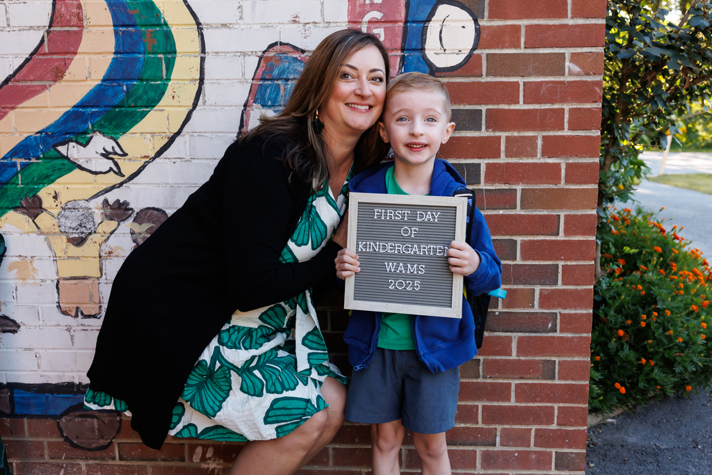 A mom and her child stand together for a photo. The child is holding a sign that says "First Day of Kindergarten, WAMS, 2025"