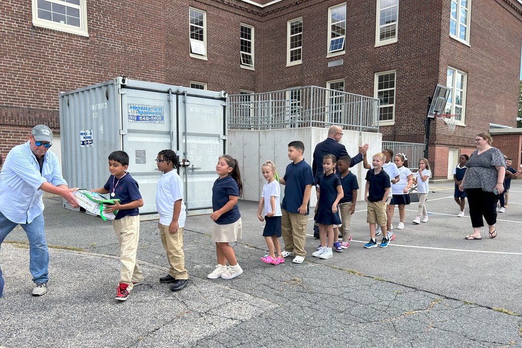A group of students line up outside a brick building, receiving gifts.