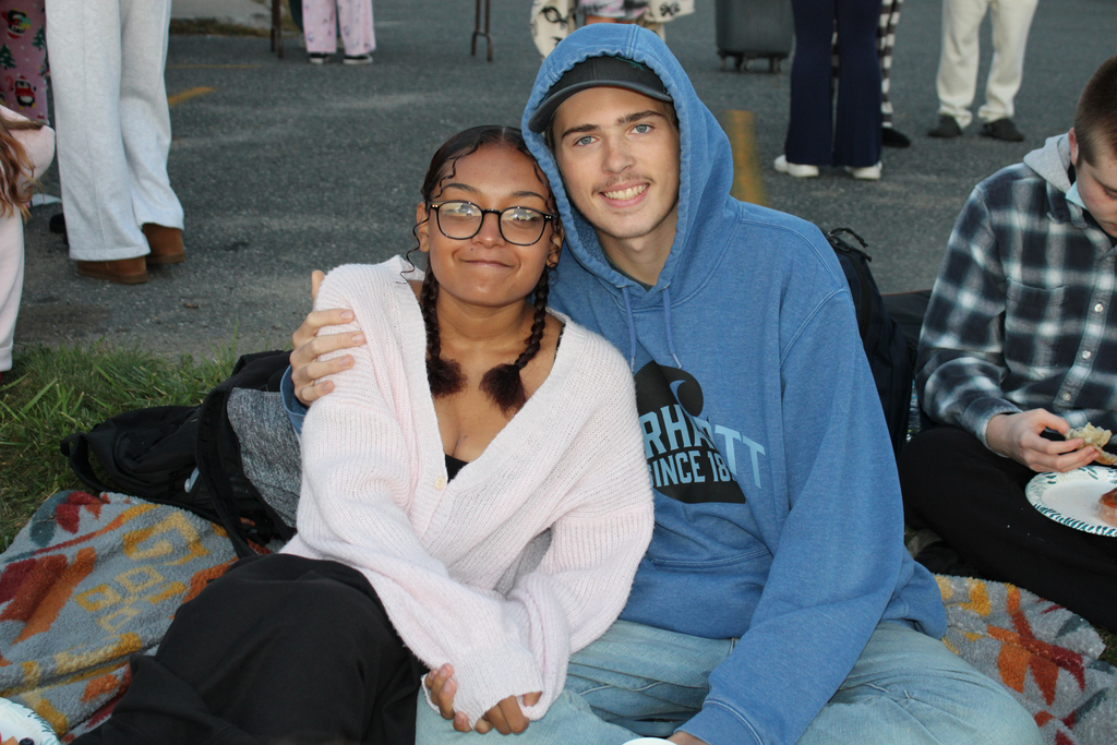 Two young people smile while sitting together outdoors, one with an arm around the other.