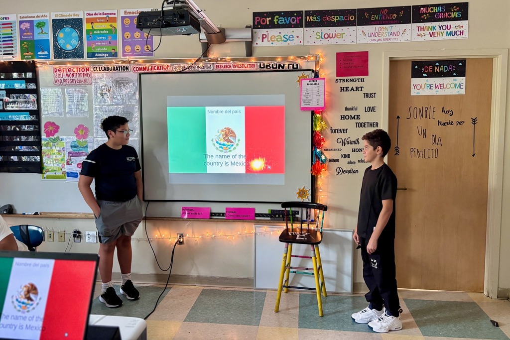 Two students stand in a classroom, one presenting a slide about Mexico's flag.