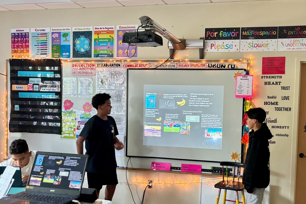 Students present a project in a classroom, with a presentation on a screen.