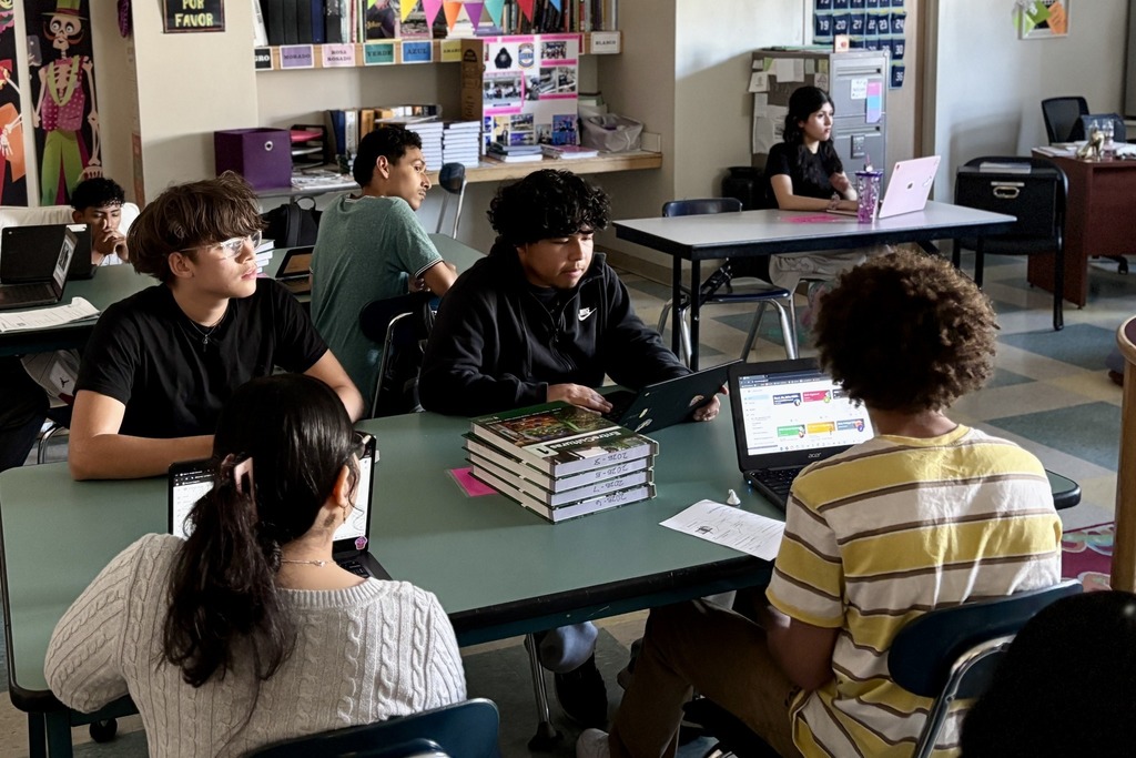 Students work on laptops in a classroom setting, some with books nearby.