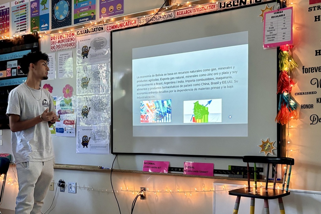 A young man presents in a classroom, with a presentation on a screen behind him.