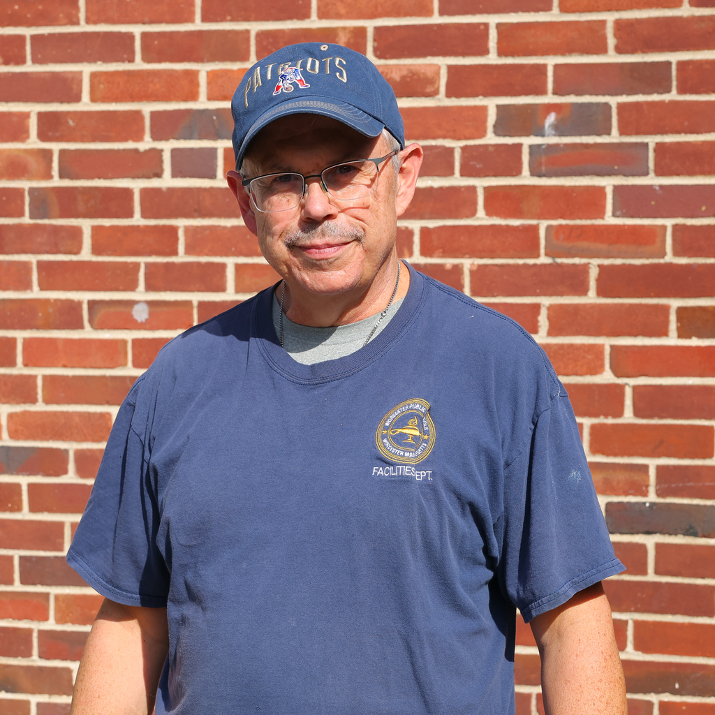 A man wearing a Patriots cap and glasses stands in front of a brick wall.