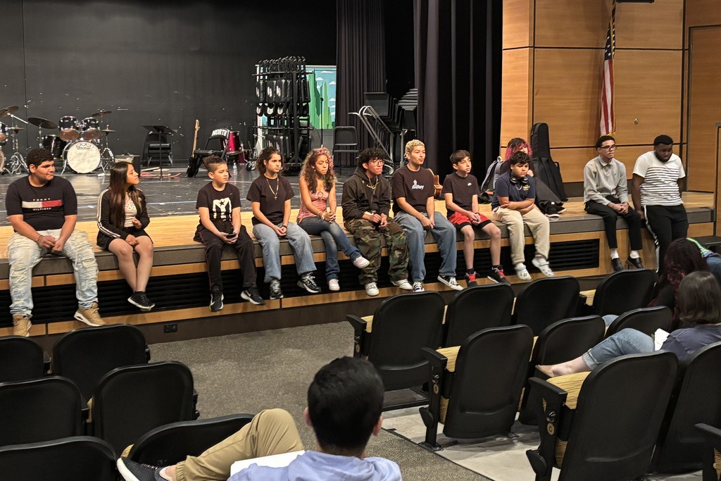 A group of diverse students sit on a stage in an auditorium.