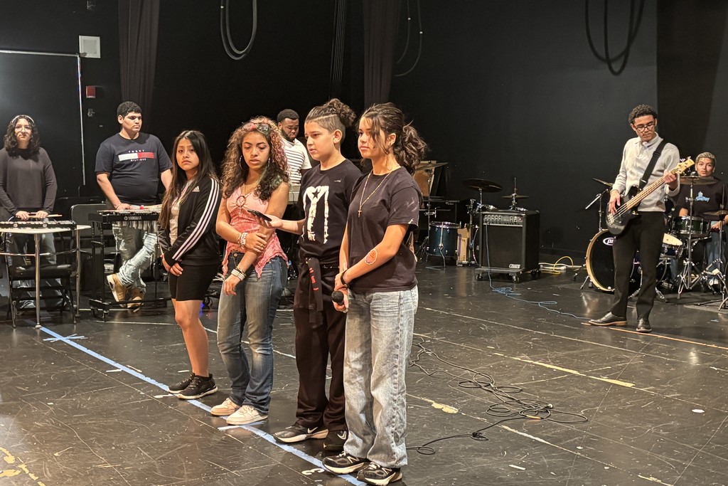 A group of young people stand on a stage, some holding microphones, with a band in the background.