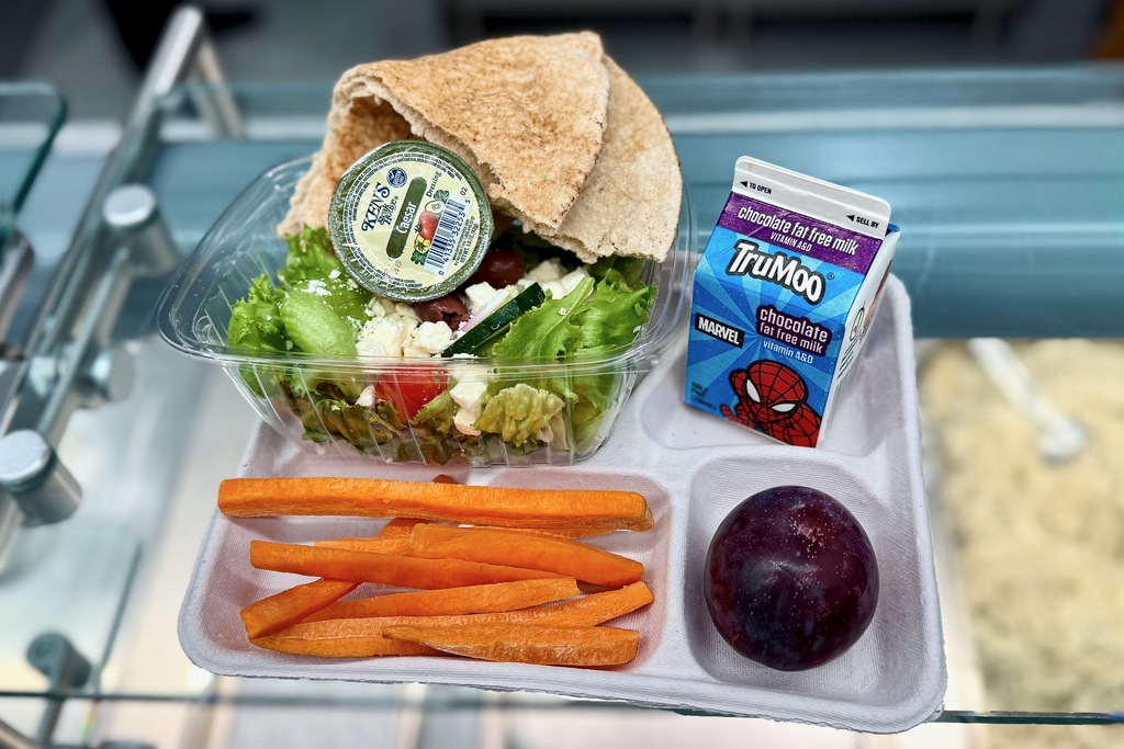 A school lunch tray with a salad, carrots, a plum, pita bread, and chocolate milk.