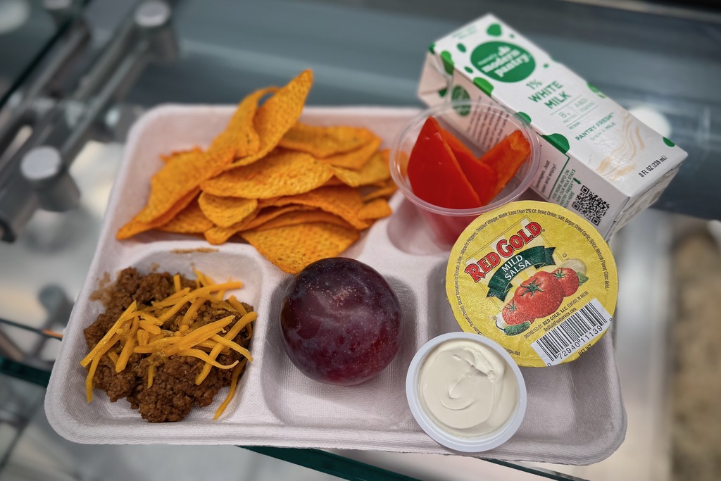 A school lunch tray with nachos, meat, cheese, salsa, milk, a plum, and peppers.