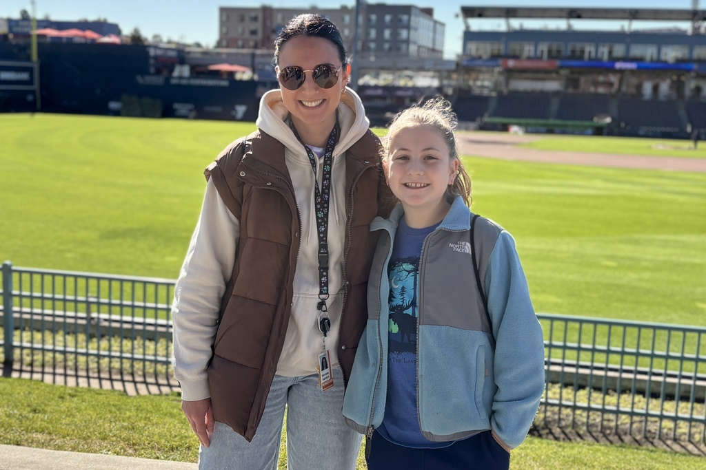 Two smiling people pose for a photo in front of a baseball field.