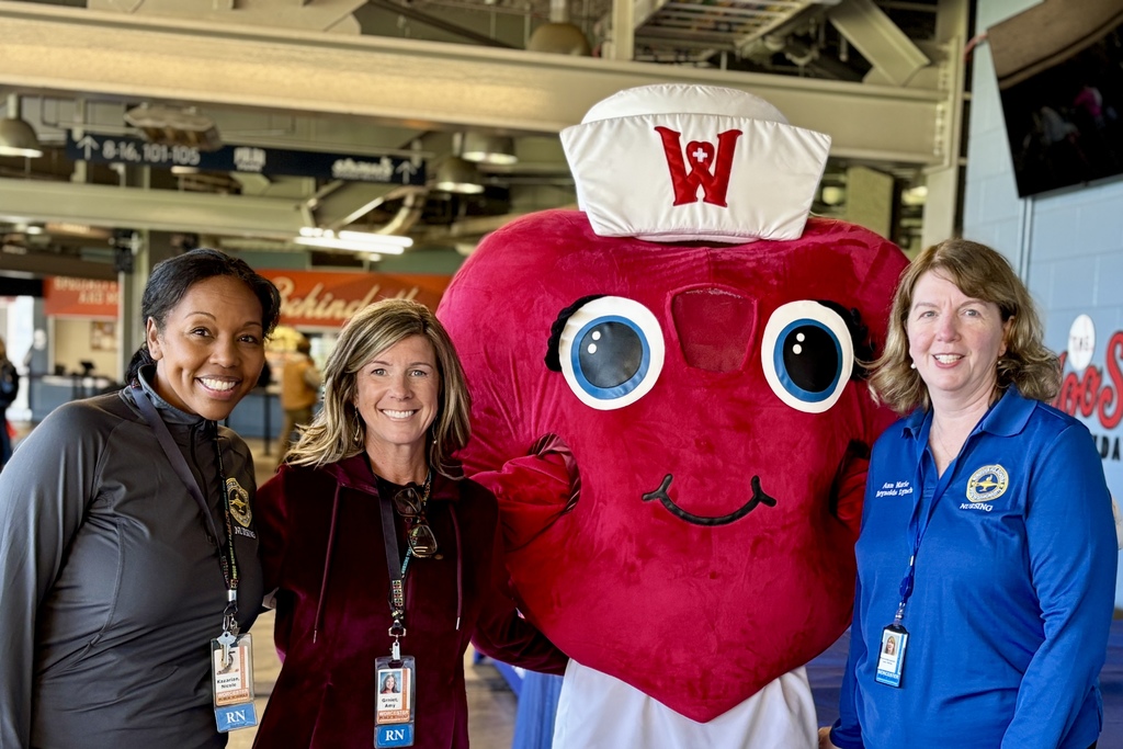 Three women pose with a heart-shaped mascot wearing a nurse's hat.