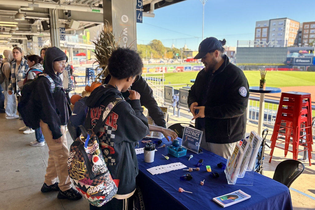 People gather at an outdoor information table with a baseball field in the background.